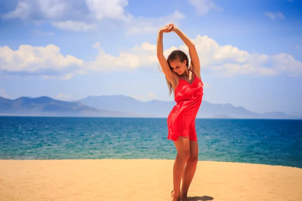 Blond girl in red stands on sand lifts hands over head