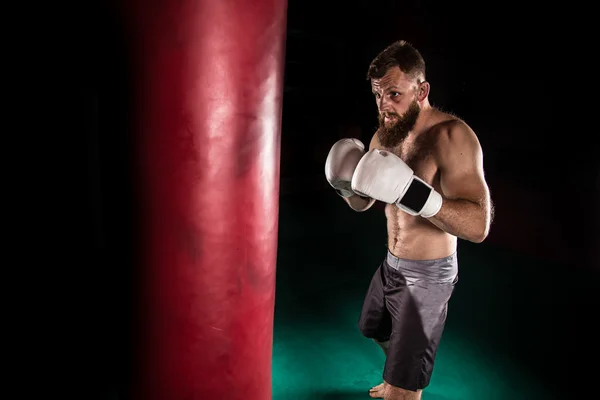 Muscular hipster fighter giving a forceful kick during a practise with a boxing bag.