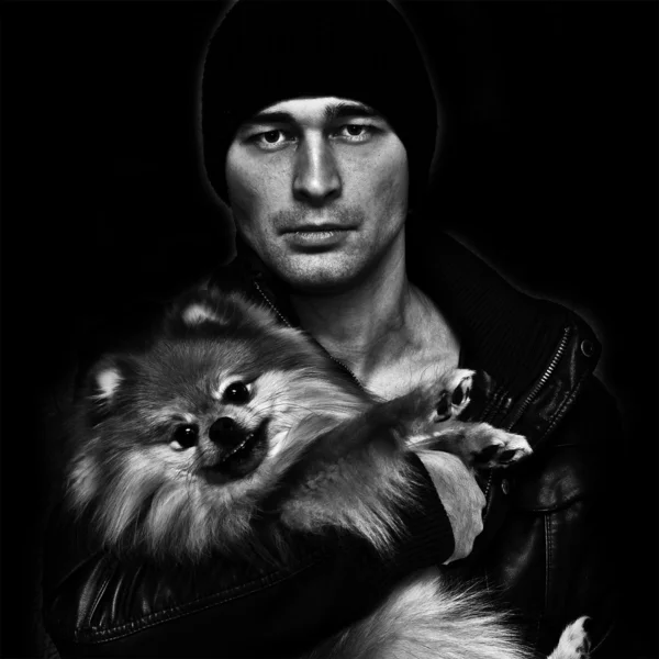 Brutal man in a hat and leather jacket with a dog in her arms on a black background