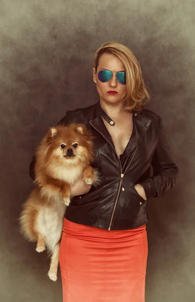 Fashionable woman in a leather jacket, sunglasses and a red skirt keeps the dog in her arms.