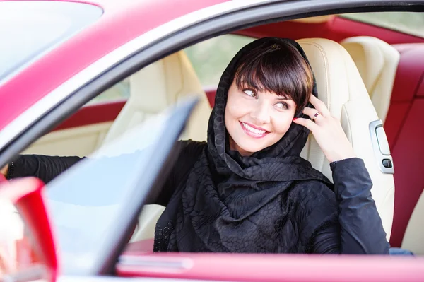 Arabic way dressed yang woman posing in red car in desert.