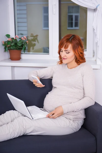 Young pregnant woman using laptop and smart phone in living room