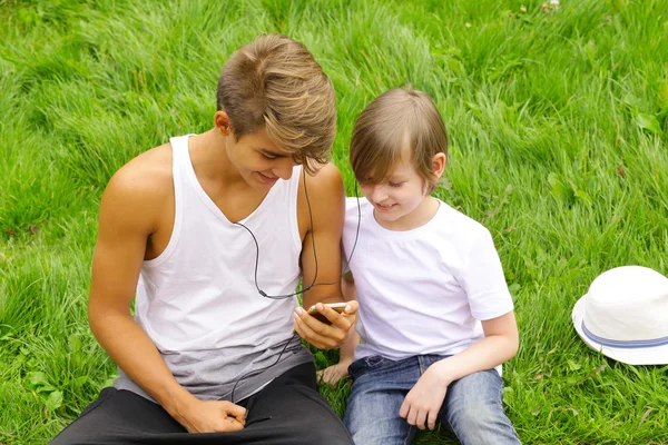 Older and younger brother sitting on the grass and looking to the smartphone