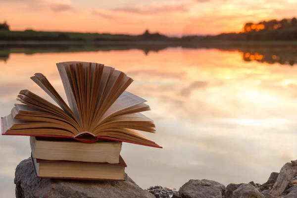 Stack of books and Open hardback book on blurred nature landscape backdrop against sunset sky with light. Copy space, back to school. Education background.