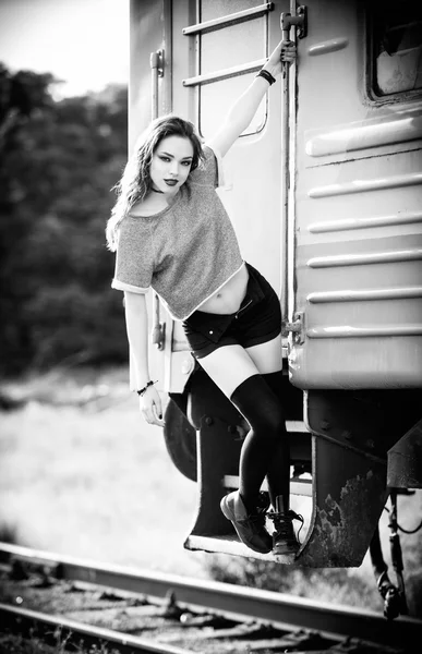 Outdoor shot: smiling young rock girl in shorts, shirt and gaiters stands near train wagon. Black and white