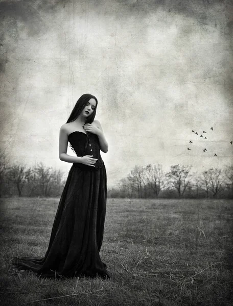 Beautiful sad goth girl stands in autumnal field. Grunge texture effect