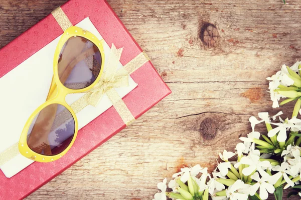 Gift box and sunglasses lie on natural wooden background with white flowers in the corner