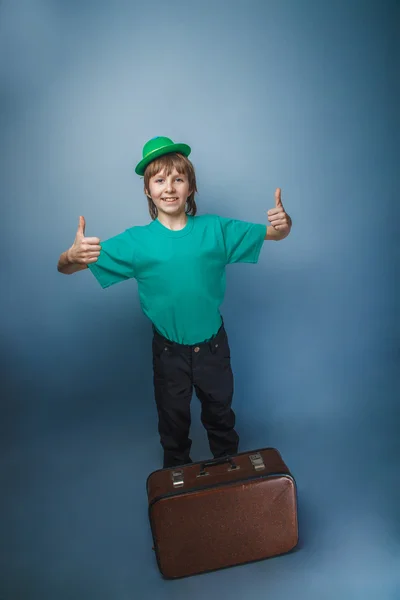 European-looking boy of  ten years in a hat holding a suitcase in hand, itinerary on a blue background