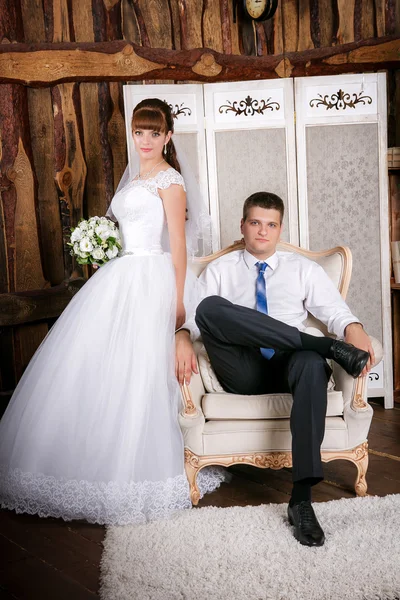 The groom sits in a chair and the  bride stands near groom in the room with a beautiful interior