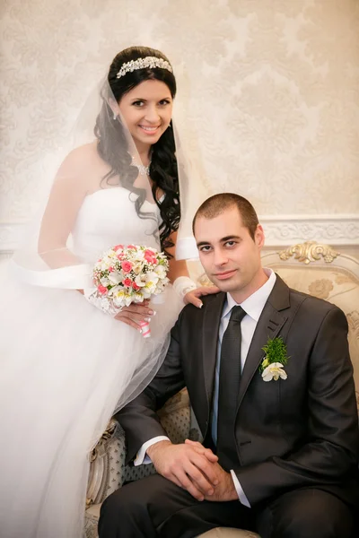 The groom sits in a chair and the bride stands near groom in the room with a beautiful interior