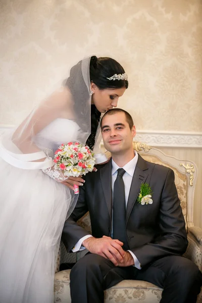 The groom sits in a chair and the bride stands near groom and kiss him in the room with a beautiful interior