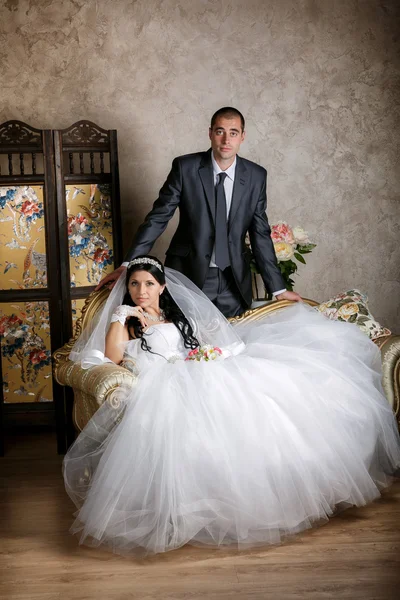 The bride sits in a chair and the groom stands near groom in the room with a beautiful interior