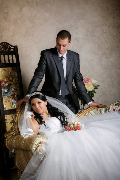 The bride sits in a chair and the groom stands near groom in the room with a beautiful interior