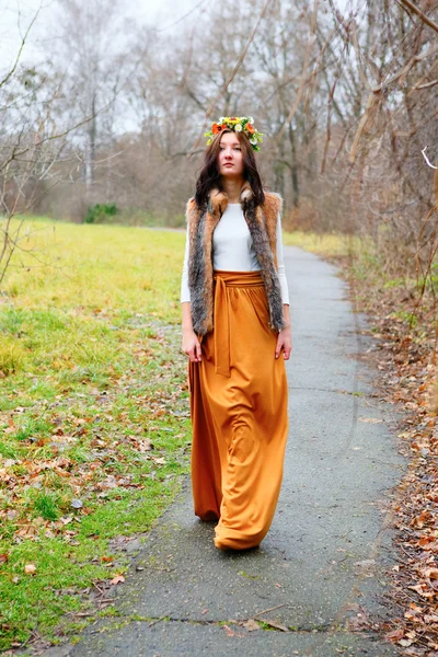 Young woman with flowers wreath goes along the asphalt path in a park