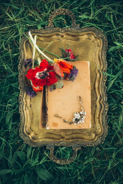 The beige book is lying on the metal bronze tray with flowers. The tray is lying on the grass. Boho style.