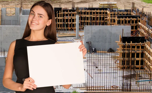 Beautiful woman smiling and hold empty paper sheet. Construction site as backdrop