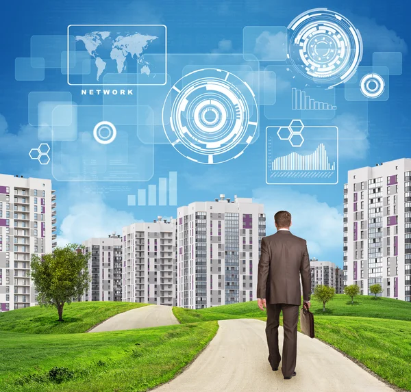 Businessman walking along road through green hills. City of tall buildings as backdrop. Charts and other virtual items in sky
