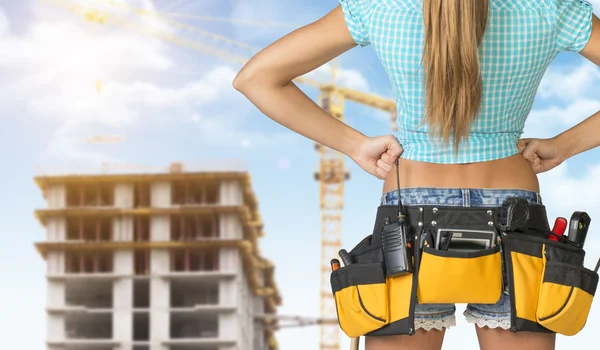 Woman in tool belt standing backwards, akimbo. Cropped image. Building under construction as backdrop