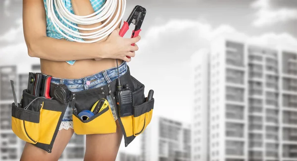 Woman in tool belt holding cable and pliers. Buildings with gray sky as backdrop