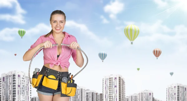 Woman in tool belt connects two flexible hoses. Buildings with air balloons as backdrop