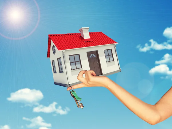 White house and keys in hand with red roof, brown door, chimney. Background sun shines brightly