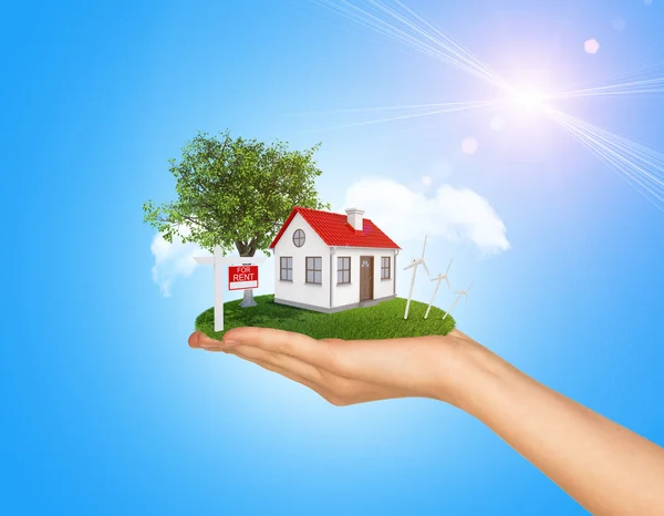 Hand holding house on green grass with red roof, chimney, tree, wind turbine. Near there is signboard for rent. Background clouds, blue sky