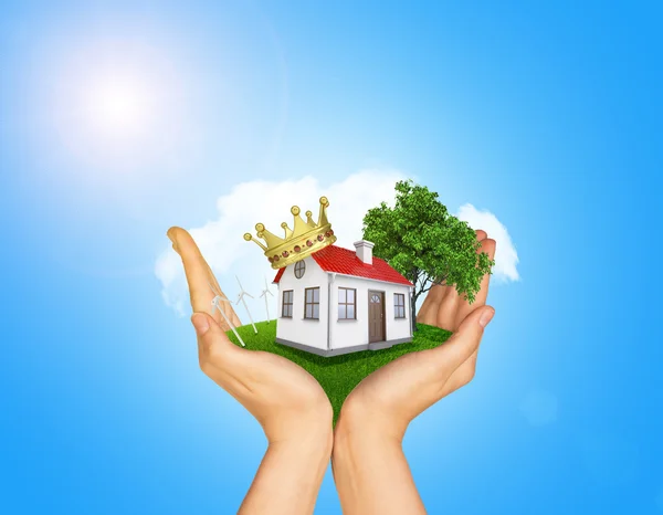 Hands holding house on green grass with crown, red roof, chimney, tree, wind turbine. Background clouds, blue sky