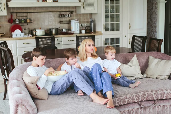 Mother and sons are sitting on the couch watching television with popcorn in the kitchen