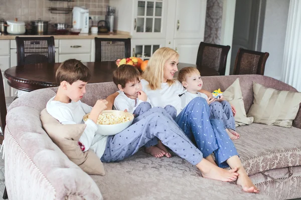 Mother and sons are sitting on the couch watching television with popcorn in the kitchen