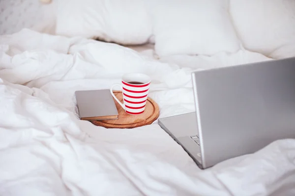 Open laptop with a cup of tea and a book lying on a white bed