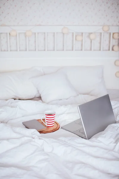 Open laptop with a cup of tea and a book lying on a white bed