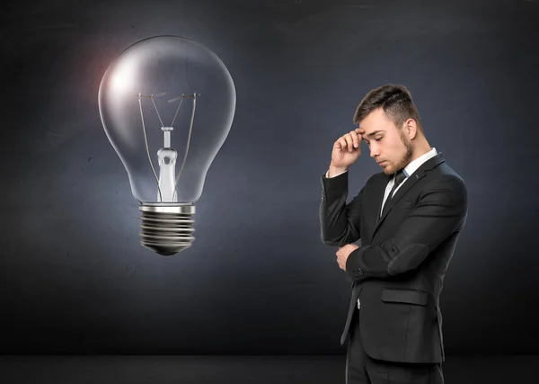 Young man in business suit thinking, on concrete wall background. Broken light bulb. Business concept