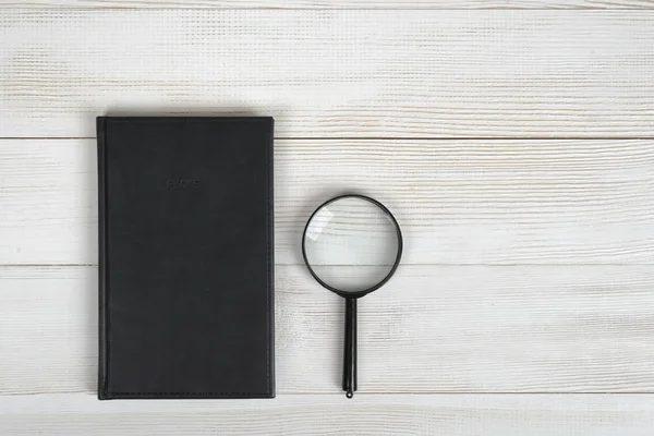 Flat lay of black notebook and magnifying glass on light wooden surface.