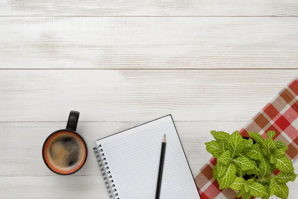 Office workplace with green houseplant on checkered tablecloth, cup of coffee and empty notebook.