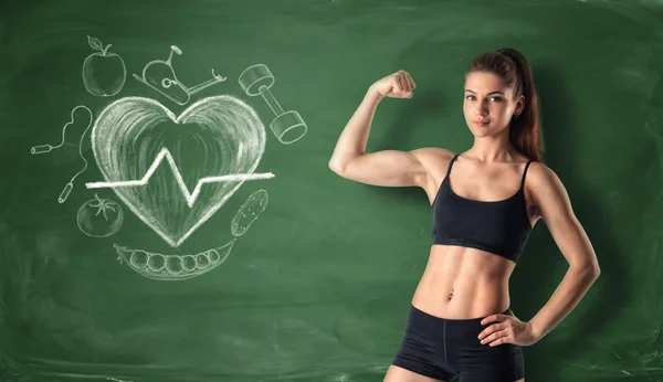 Fitness girl showing her bicep on the background of a chalkboard with drawn beating heart and sport doodles