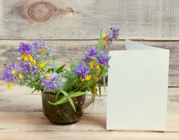 Cup and a bouquet of wild flowers and a blank sheet of paper on a wooden surface.