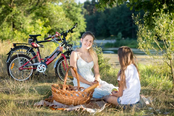 Smiling woman sitting on blanket under big tree with her daughte