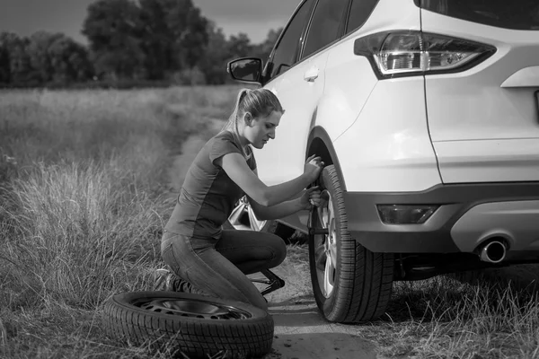 Black and white image of upset young woman changing car wheel on