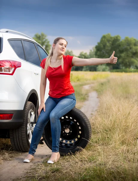 Beautiful woman sitting at broken car and hitchhiking at field