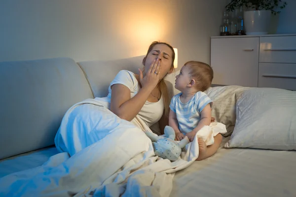 Baby boy sitting on bed and looking at yawning tired mother