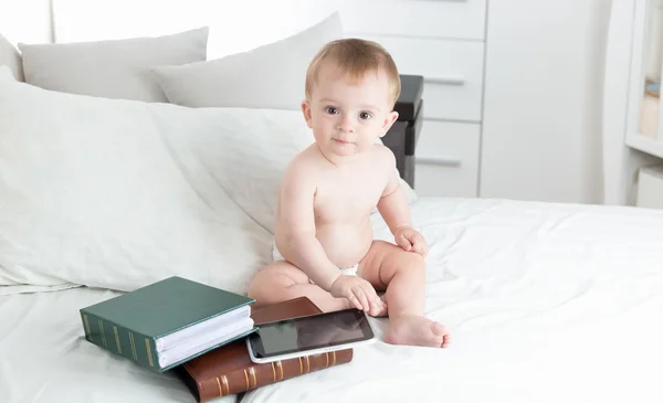Cute naked baby in diapers with books and digital tablet on bed