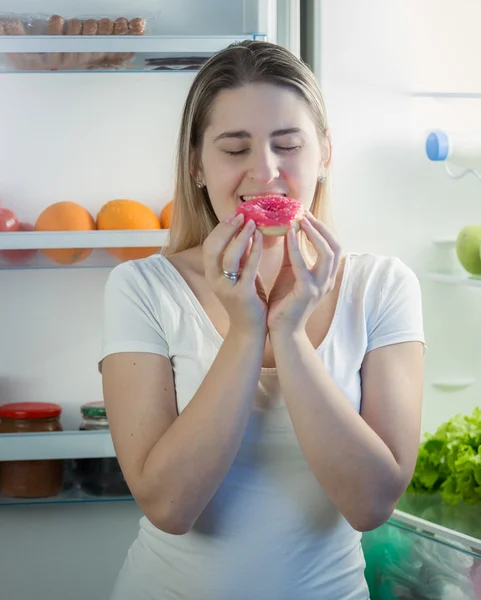 Young woman eating big donut on kitchen at night. Concept of unh