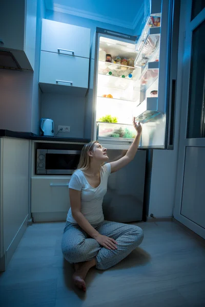 Woman sitting on floor and reaching for food from open refrigera