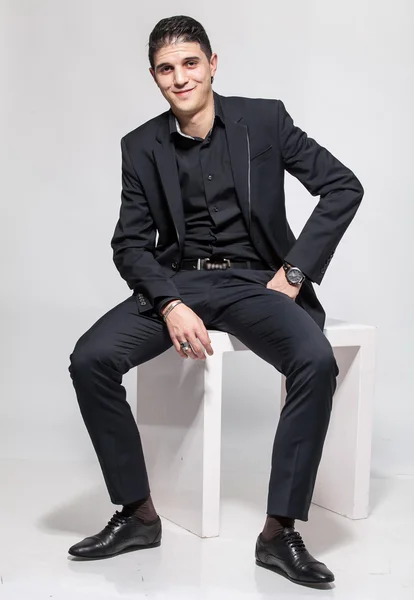 Latin man in black suit sitting on white chair and smiling