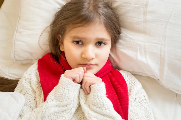 Little sad girl in white sweater lying under blanket at bed