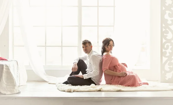 Pregnant couple sitting back to back on floor against big window
