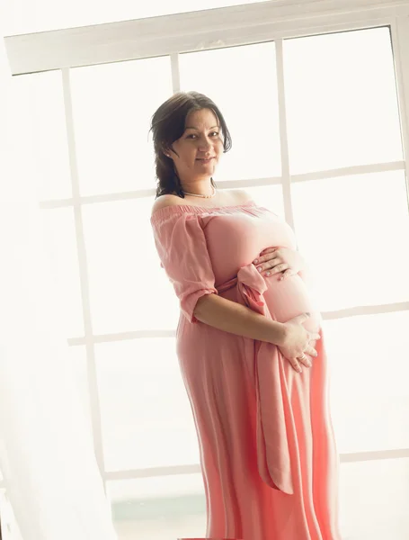 Toned shot of pregnant brunette woman posing against big window