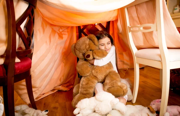 Smiling girl in pajamas hugging teddy bear at self-made house