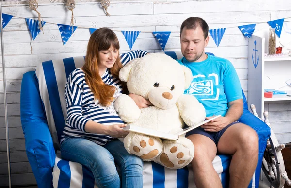 Pregnant woman and husband reading big book to teddy bear on sof