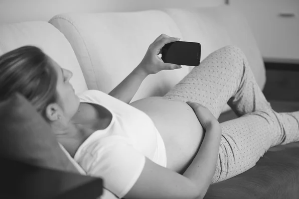 Black and white shot of pregnant woman relaxing on sofa and usin